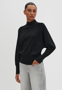 Pullover nero con collo alto, maniche lunghe a raglan, vita aderente e texture morbida; abbinato a jeans grigi chiari a gamba larga.