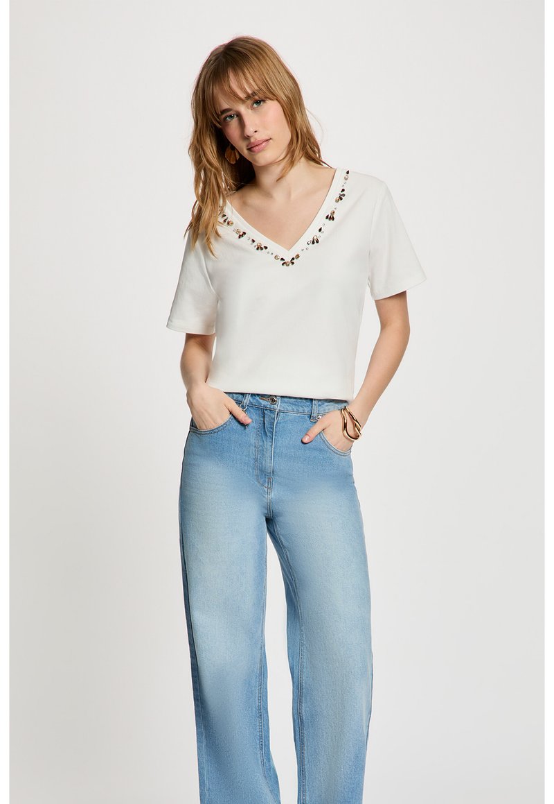 Woman with light brown hair wears a white V-neck top with beaded trim and light blue high-waisted wide-leg jeans, hands in pockets.
