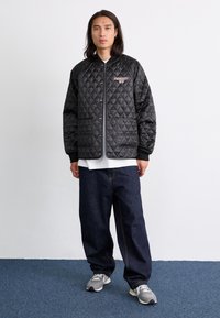 Sort quiltet jakke med diamantmønster, trykknapper, ribbede manchetter og to forlommer, båret over en hvid skjorte og mørkeblå jeans.
