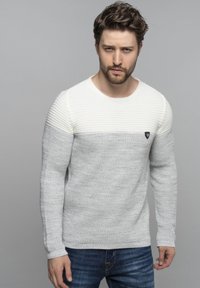 Grijs en wit gebreide trui met ribbelstructuur op de schouders, aansluitende mouwen en een klein logo-accent op de borst.