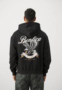 Crna dukserica s velikom grafikom orla i natpisom "Benfica" u bijeloj i zlatnoj boji. Mehak materijal, prednji džep i kapuljača s podesivim vezicama.