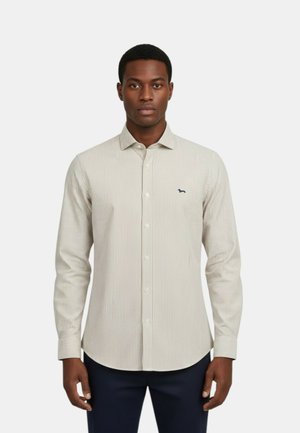 Uomo che indossa una camicia beige a righe verticali con piccoli bottoni e un piccolo logo scuro sul petto, in piedi davanti a uno sfondo chiaro uniforme.