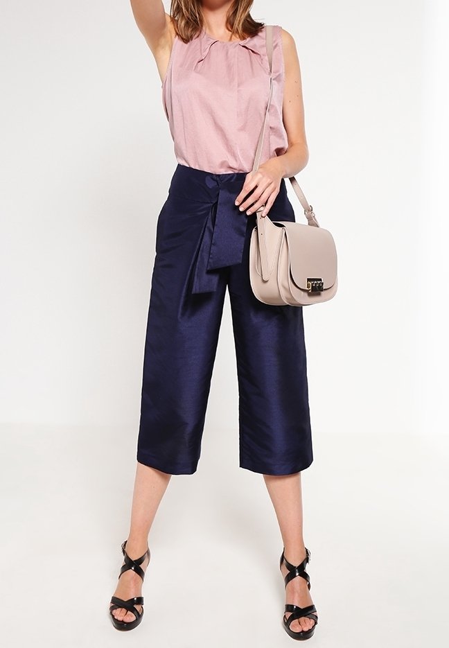 Top senza maniche di un leggero rosa con dettaglio a fiocco, abbinato a pantaloni cropped in satin blu navy e una borsa a tracolla beige. Sandali neri con cinturini completano il look.