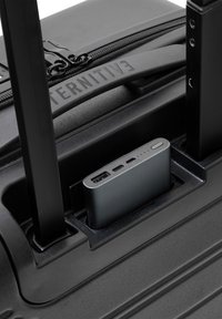 Schwarzer Koffer mit strukturiertem Finish, ausgestattet mit einer integrierten grauen Powerbank, die im Griffbereich eingesetzt ist, mit sichtbaren USB-Anschlüssen.