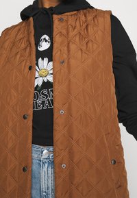 Hnědá prošívaná vesta s diamantovým vzorem a patentními knoflíky, nošená přes černou mikinu s květinovou grafikou. Na spodní části jsou vidět modré džíny.