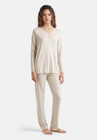 Beige sada loungewear, která zahrnuje dlouhý rukáv top s našitým krajkovým výstřihem ve tvaru vln a odpovídající kalhoty s krajkovými detaily na lemu. Měkká textura.