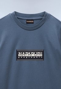 NapaPuri graafiline t-särk sinisest puuvillast ümarate krae. Keskosas on must logo silmapatch valgete kirjade ja aktsentidega.