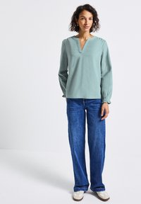 Femme aux cheveux bouclés portant un blouse à col en V à manches longues de couleur vert clair, un jean bleu et des baskets blanches devant un fond blanc.