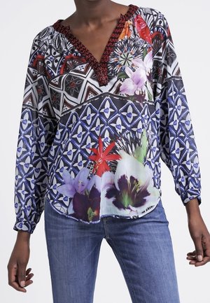 Femme portant un chemisier à manches longues avec des motifs géométriques et floraux en noir, blanc, rouge, violet et vert, associé à un jean bleu.