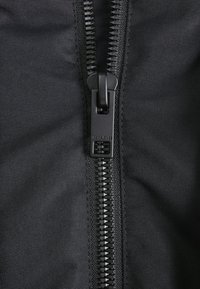 Fermeture éclair noire sur un tissu texturé, avec des dents dentelées et une tirette. La fermeture éclair est centrée et fonctionnelle, avec le nom de la marque visible.