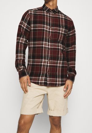 Camisa de franela a cuadros burdeos con cierre de botones y mangas largas, combinada con pantalones cortos beige enrollados, mostrando un ajuste relajado.