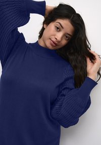 Maglione blu navy con collo alto e maniche a sbuffo a coste. Texture morbida con un corpo aderente e maniche voluminose e testurizzate.