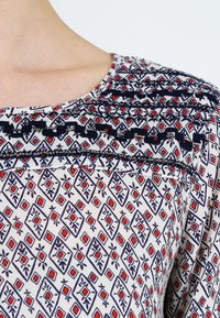 Blouse en tissu blanc présentant un motif géométrique en bleu marine, rouge et noir. Des coutures décoratives le long de l'encolure ajoutent de la texture.