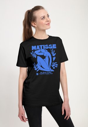 Femme avec une queue de cheval portant un T-shirt noir avec une œuvre bleue de Matisse et un texte, debout devant un fond clair uni.
