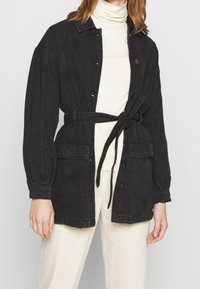 Giacca di jeans nera con grandi tasche frontali, coulisse in vita, chiusura con bottoni e maniche lunghe. Indossata sopra un dolcevita crema.