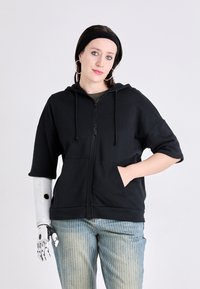 Crna zip-up dukserica s opuštenim krojem, dva prednja džepa i vezicama. Nosite je s svijetloplavim trakom i bijelim prugastim trapericama. Vidljiv je robotski ručni mehanizam.