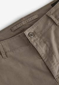 Pantaloni in cotone beige con una texture liscia, dotati di chiusura con bottone e passanti per cintura. Dettagli di cucitura discreti lungo la vita.