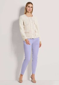 Crème tweed jas met franjes en twee voorzakken, gecombineerd met licht lavendelkleurige slim-fit broek en strap-sandalen in zilver.