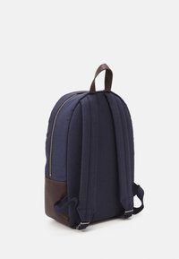 Marinblå tyg ryggsäck med brunt läderbotten, quiltad textur och justerbara remmar. Har ett bärhandtag och en silverfärgad dragkedja.