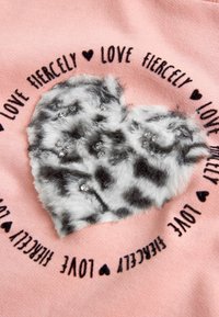 La tela rosa presenta un corazón de peluche gris y blanco con manchas negras, adornado con strass, rodeado del texto "Ama con intensidad."
