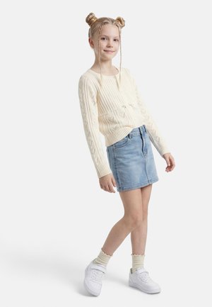 Fată cu păr blond prins în două cocuri, purtând un cardigan tricotat crem, fustă de denim albastru deschis, șosete crem și adidași albi, stând pe un fundal alb.