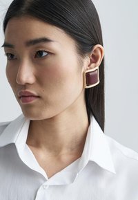 Štvorcové červené náušnice s lesklým povrchom, rámované svetlom zlatým kovom; modelka má na sebe bielu košeľu s golierom.