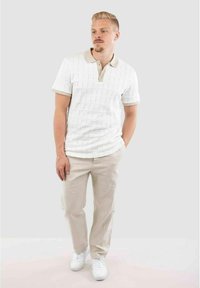 Polo shirt blanc texturé avec des manches courtes et des accents beiges, associé à un pantalon beige clair et des baskets blanches. Design simple et épuré.