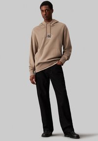Hanorac bej cu glugă, cu buzunar frontal și imprimeu grafic, asortat cu pantaloni negri cu croi larg și pantofi negri. Design simplu, fără modele.