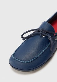 Mocassin en cuir bleu avec détail cousu et lacets noués sur fond blanc.
