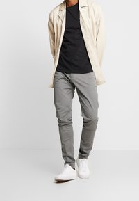 Béžová lehká bunda přes černé tričko, doplněná šedými slim-fit kalhotami a bílými teniskami. Hladká textura, minimalistický design.