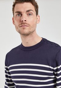 Pull marin avec des rayures horizontales blanches, texture en tricot côtelé et trois boutons sur l'épaule gauche.