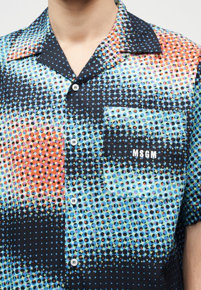 Gros plan d'une personne portant une chemise à manches courtes multicolore à pois, avec une poche avant étiquetée "MSGM" et des boutons blancs.