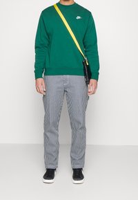 Grön sweatshirt med rundad halsringning och vit logotyp, randiga grå byxor, svarta sneakers och en axelväska med gul rem.