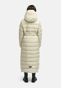 Lång, puffy, lys beige vinterjakke med indsnævret talje og hætte. Har horisontale syninger og et logo mærke nær kanten.
