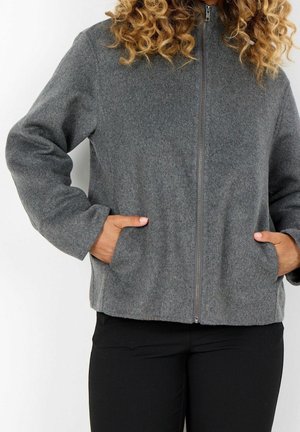 Person mit lockigem Haar, trägt eine graue Jacke mit Reißverschluss und hat die Hände in den Seitentaschen, kombiniert mit schwarzen Hosen.