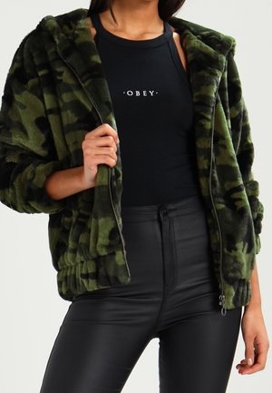 Person trägt eine grüne Tarnmuster-Kunstpelzjacke über einem schwarzen ärmellosen Oberteil mit dem Schriftzug "OBEY" und schwarze hoch taillierte Hosen.