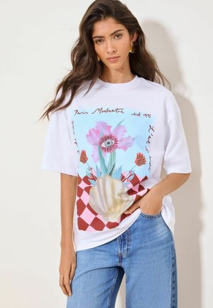 Tânără femeie purtând un tricou alb supradimensionat cu un grafic floral și scoică suprarealistă, băgat în blugi albaștri, stând în fața unui fundal simplu.