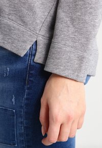 Felpa grigia chiara con un design cropped, polsini a costine e fessure laterali, abbinata a jeans scuri strappati. Mano appoggiata sui jeans.