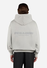 Sudadera gris claro con una gran impresión en la parte posterior que dice "PEGADOR" y "CERTIFICADO AUTÉNTICO DE STREETWEAR". Corte holgado con puños de canalé.