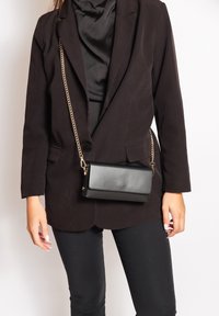 Blazer noir sur un haut noir, associé à un petit sac à main noir avec une chaîne dorée. Le sac a une texture lisse et un design minimal.