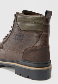 Smeđe kožne čizme s maslinasto zelenim naglaskom, koje se odlikuju teksturiranim materijalima, kontrastnim šavovima i izdržljivom gumenom đonom. Prisutno Wrangler logo.
