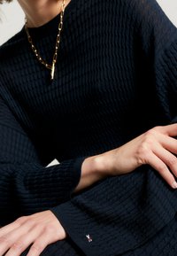 Suéter de punto azul marino con un patrón texturizado, mangas anchas y un pequeño logo bordado en el dobladillo. Collar de cadena dorada que realza el look.