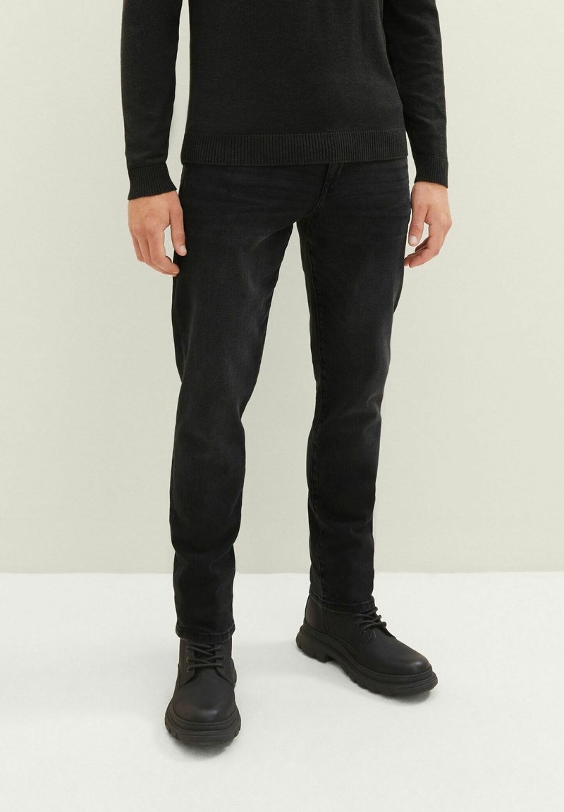 Schwarze Jeans mit slim fit und subtiler Textur, kombiniert mit schwarzen Schnürstiefeln. Der Pullover ist schwarz mit gerippten Bündchen und Saum.