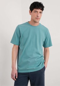 Turkos bomullst-shirt med rund hals, korta ärmar och avslappnad passform, kombinerad med mörkblå byxor, mot en neutral bakgrund.