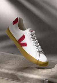 Baltie sneakers ar sarkaniem akcentiem, ar teksturētu gumijas zoli, auklu aizdari un lielu "V" logotipu sānos un uz papēža.