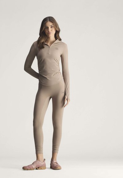 Young woman stands wearing beige long-sleeve top and leggings with pink minimalist shoes on plain light background.