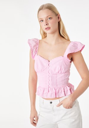 Joven mujer con una blusa sin mangas rosa con volantes y botones, combinada con pantalones blancos, posando frente a un fondo blanco.