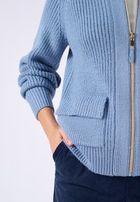 Cárdigan de punto azul claro con cremallera frontal, textura acanalada y un bolsillo delantero. Combinado con pantalones de pana oscuros.