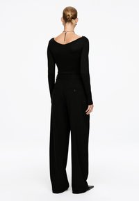 Frau mit einem Dutt, die ein schwarzes langärmliges Oberteil und hochtaillierte, weite schwarze Hosen trägt, steht mit dem Rücken zu einem weißen Hintergrund.