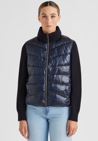 Navyblauer Steppwesten mit hohem Kragen, Reißverschluss und horizontalem Steppmuster; gerippte schwarze Ärmel.